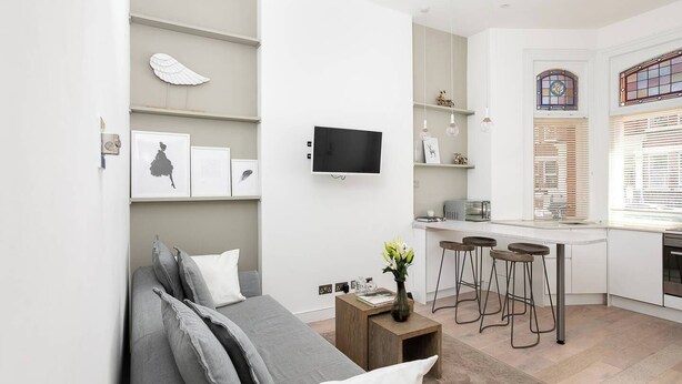 Ein modernes Wohnzimmer mit grauem Sofa, einem Couchtisch und einem an der Wand befestigten Fernseher. Es gibt eine Küchenzeile mit einem Barhocker und einem Fenster mit Jalousien.