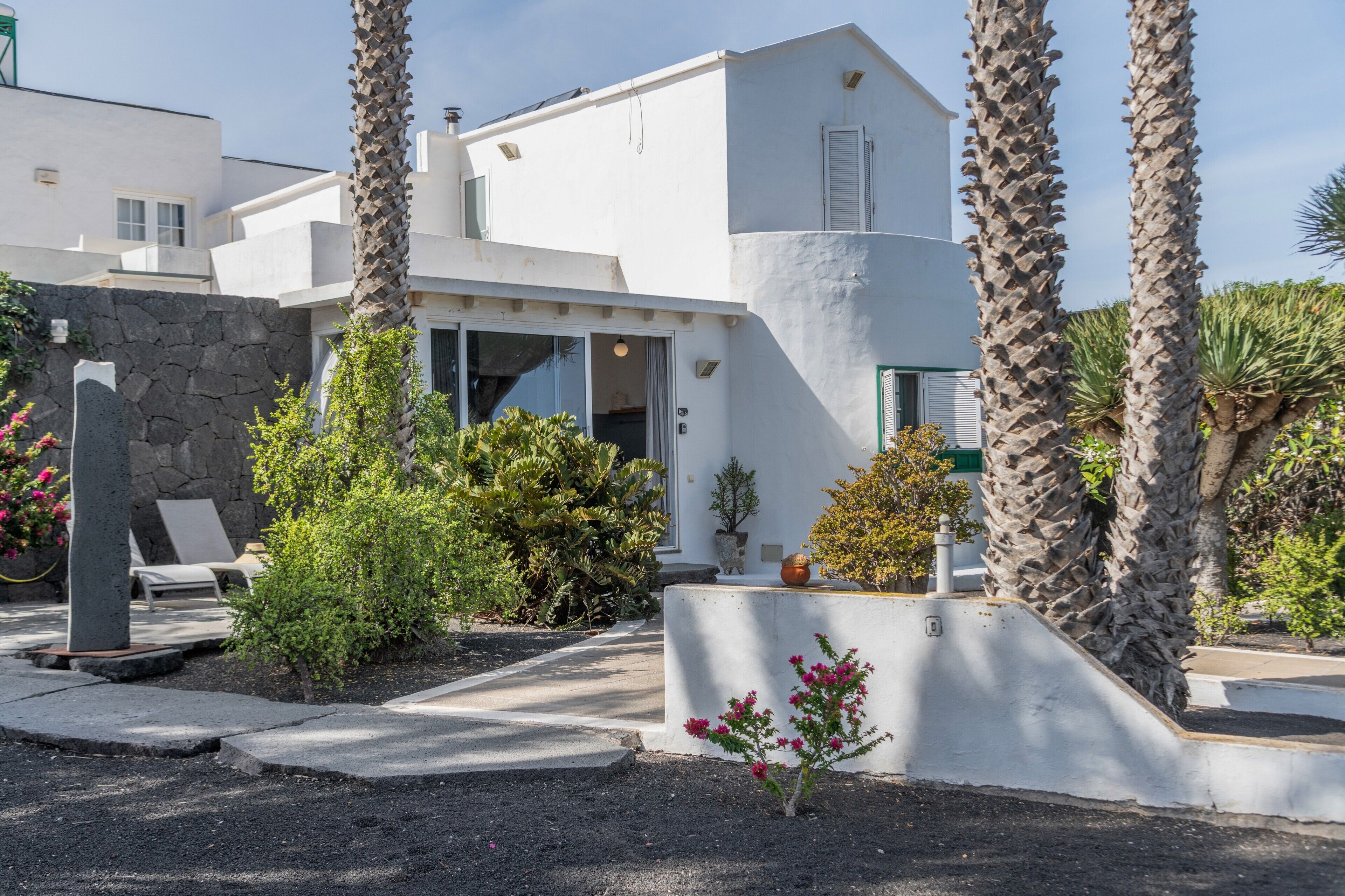 A modern white building with a stone wall, palm trees, and a small garden with plants and flowers.