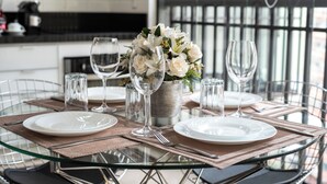 Un tavolo da pranzo rotondo in vetro con piatti bianchi, calici di vino trasparenti e un centrotavola con fiori bianchi in un cesto intrecciato.