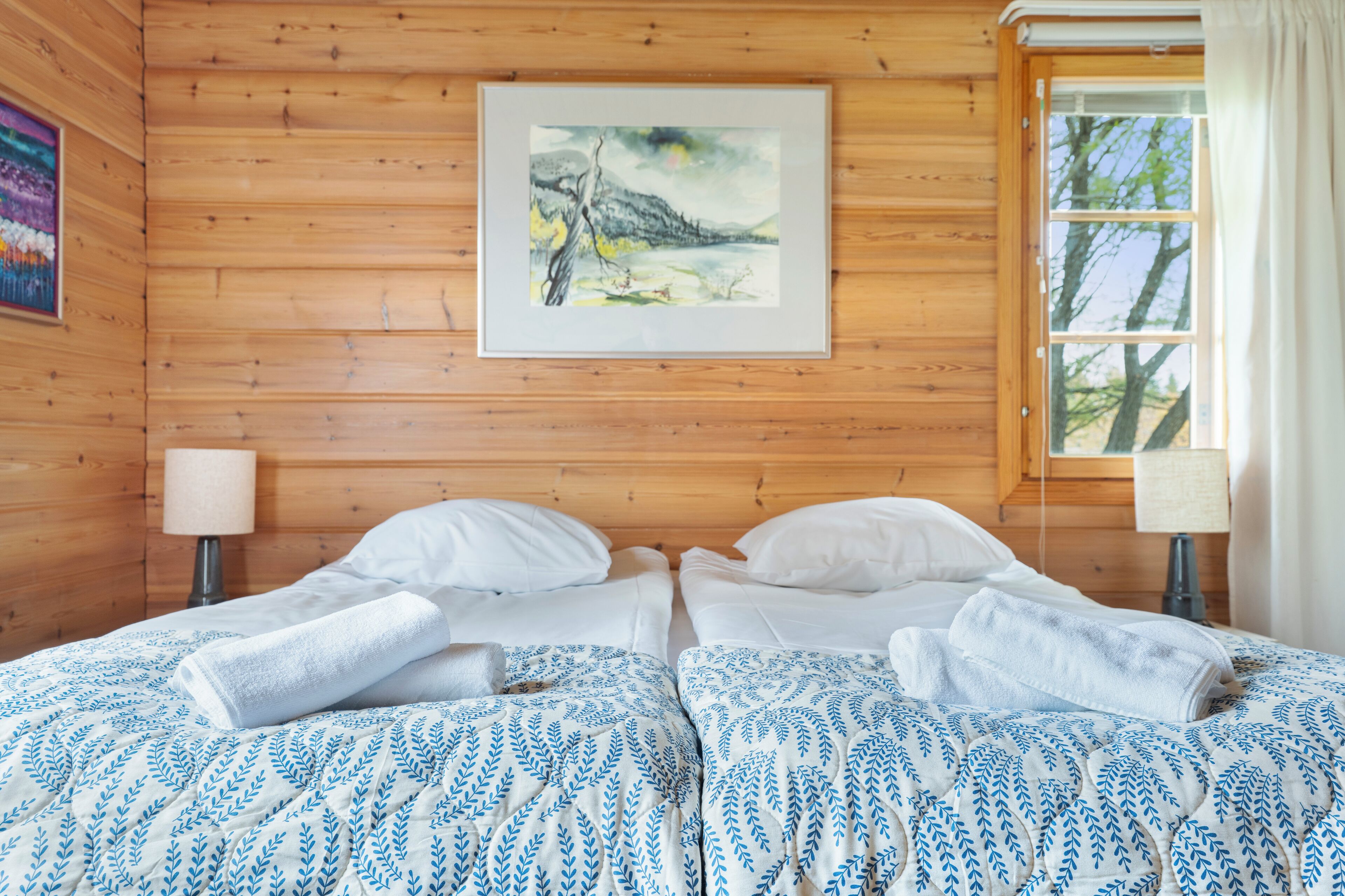 Habitación de cabaña de madera con una cama, dos lámparas de noche, un cuadro en la pared y una ventana con cortinas.