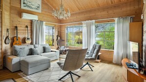 A cozy cabin interior with a wooden ceiling, a chandelier, a grey sofa, a grey armchair, and a dining table.