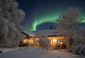 Une maison recouverte de neige, dotée d’un design moderne, entourée d’arbres, sous un ciel nocturne illuminé par des aurores boréales.