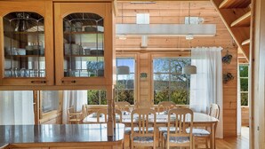 A wooden dining room with a large table and chairs, a kitchen area with wooden cabinets, and large windows with curtains.