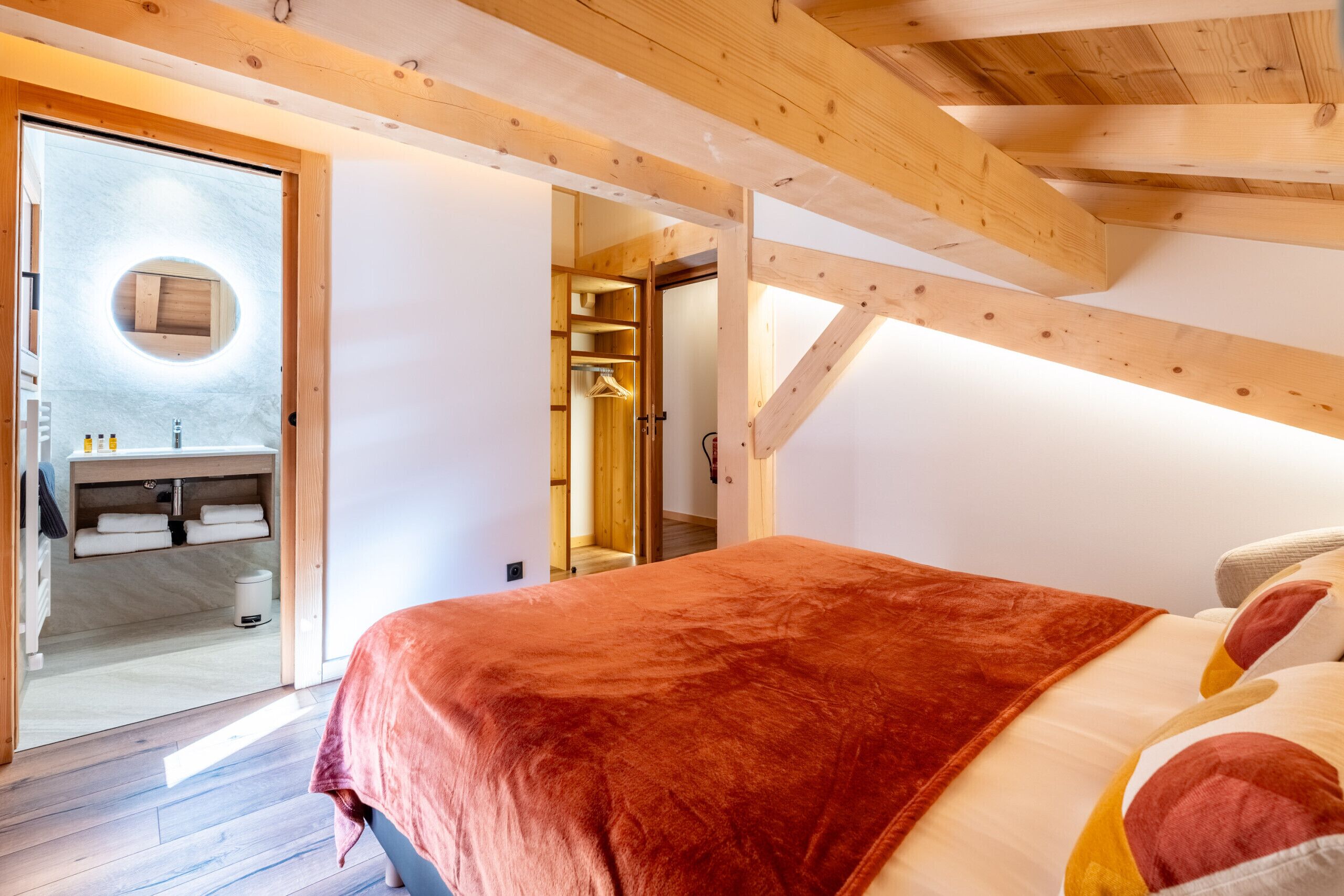 A bedroom with a wooden ceiling, a bed with a red blanket, a circular mirror, and a bathroom with a sink and towels.