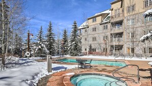 Un espace piscine avec un jacuzzi, entouré d’arbres couverts de neige et d’un bâtiment doté de balcons.
