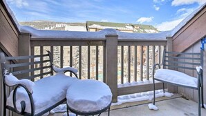 Un balcon avec deux chaises en métal recouvertes de neige, une clôture en bois et une vue sur un paysage enneigé avec des bâtiments en arrière-plan.