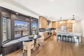 Une cuisine moderne avec des meubles en bois, un îlot central doté de tabourets de bar et de grandes fenêtres offrant une vue sur les montagnes.
