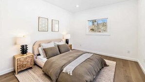 Une chambre à coucher avec un lit, deux tables de chevet, une fenêtre et deux tableaux encadrés accrochés au mur.