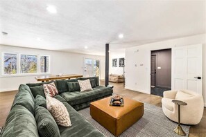 A modern living room with a sectional sofa, a coffee table, and a round armchair. There is a dining area with a table and chairs, and a door leading to another room.