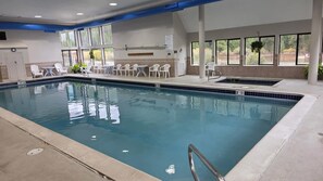 A swimming pool with a tiled deck, white lounge chairs, and a view of the outdoors through large windows.