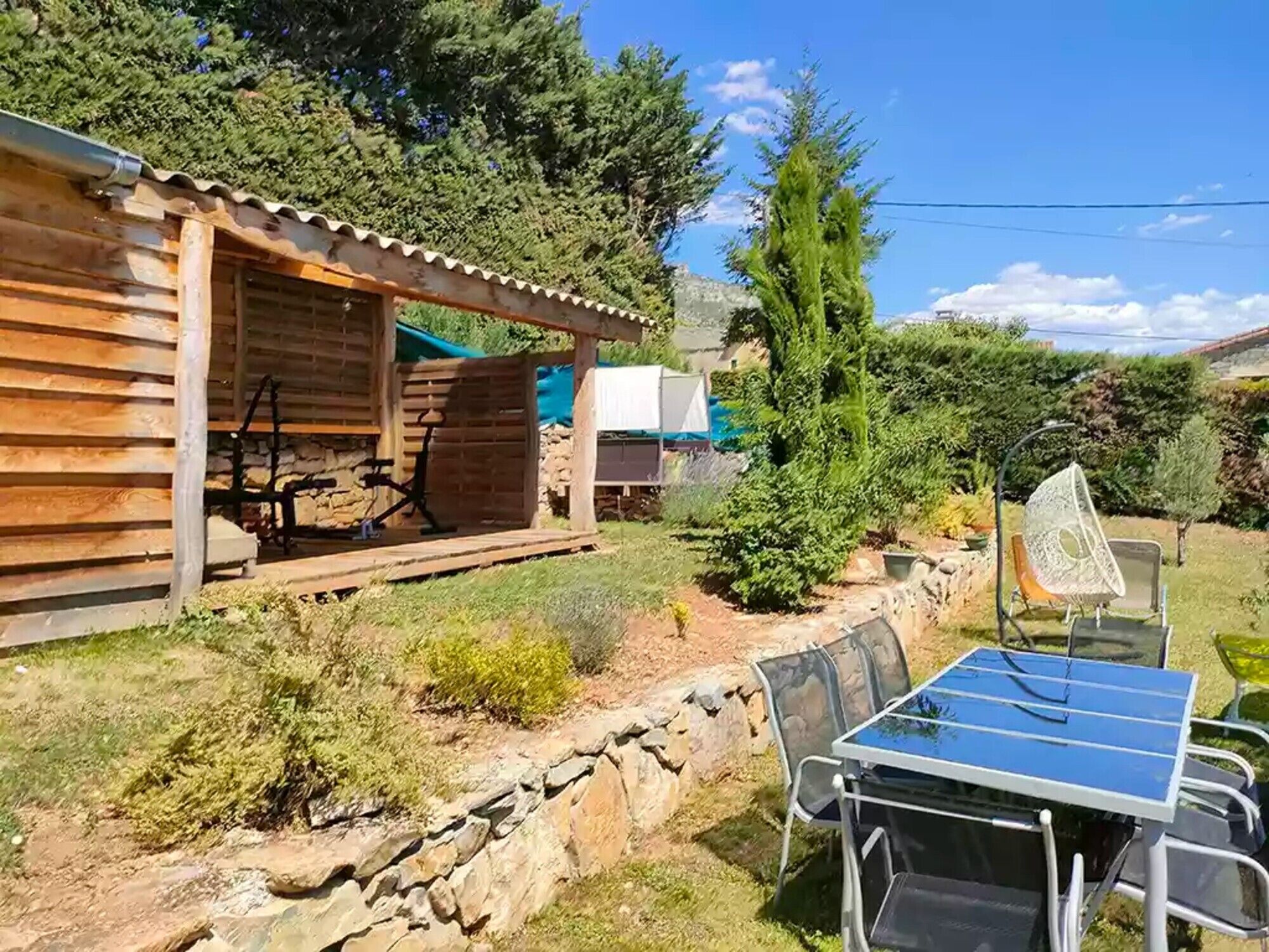 Een rustieke houten hut met een terras, een stenen muur en een zonne-energie tafel met stoelen.