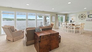 A spacious living room with a view of a lake, a dining area with white furniture, and a large wooden coffee table. - Winding Bay - Lakefront with stellar main channel views! (Sunrise Beach)