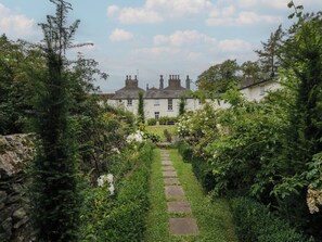 A traditional house with a stone pathway leading to the entrance, surrounded by lush greenery and flowering plants.