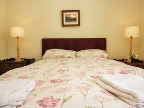 A hotel room with a large bed, two bedside lamps, a framed picture on the wall, and a nightstand with a lamp and flowers. - The Gameskeepers Cottage (Grange-over-Sands)