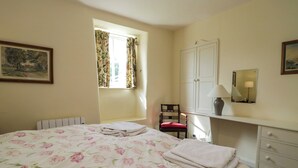 A bedroom with a large bed, a chair, a dresser, a window with curtains, and a painting on the wall. - The Gameskeepers Cottage (Grange-over-Sands)