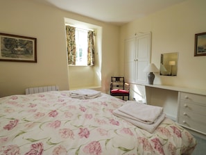 A bedroom with a large bed, a chair, a dresser, a window with curtains, and a painting on the wall. - The Gameskeepers Cottage (Grange-over-Sands)
