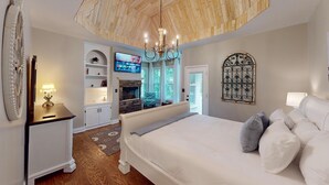 A bedroom with a large bed, a TV mounted on the wall, a fireplace, and a chandelier.