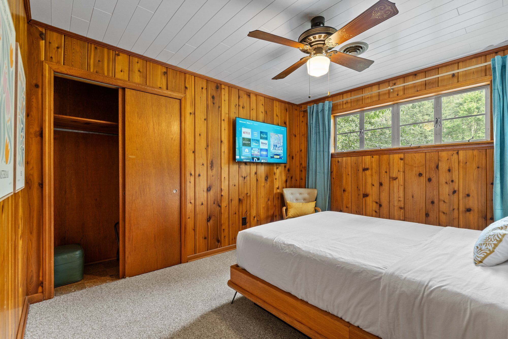 A bedroom with a wooden ceiling, a bed, a chair, a television, and a window with curtains.
