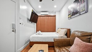 Habitación de hotel compacta con cama, sofá de cuero marrón, mesita pequeña y televisor montado.