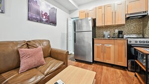 Un espacio habitable compacto con kitchenette, un sofá de cuero marrón, una pequeña mesa de madera y un cartel con un panorama urbano en la pared.
