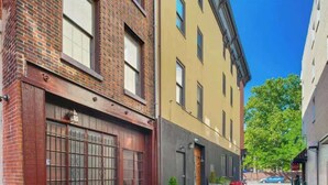 A narrow urban street with two-story buildings, one with a red brick facade and another with a yellow and gray facade. There are potted plants and a red bicycle parked on the sidewalk. - Cozy Studio Retreat in Philadelphia (Philadelphia)