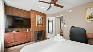 A hotel room with a brick wall, a fireplace, a bed, a desk, a chair, a television, and a ceiling fan. - Spacious Family-Friendly Home in Philadelphia (Philadelphia)