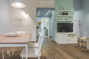 Een moderne eetruimte met een houten tafel en witte stoelen, een witte kast met een flatscreen-tv, en een gang die naar een keuken leidt.