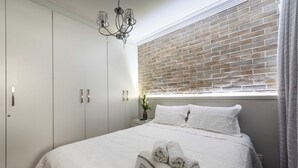 A neatly made bed with white linens and rolled towels, a small bedside table with a vase, and a brick-patterned window blind.