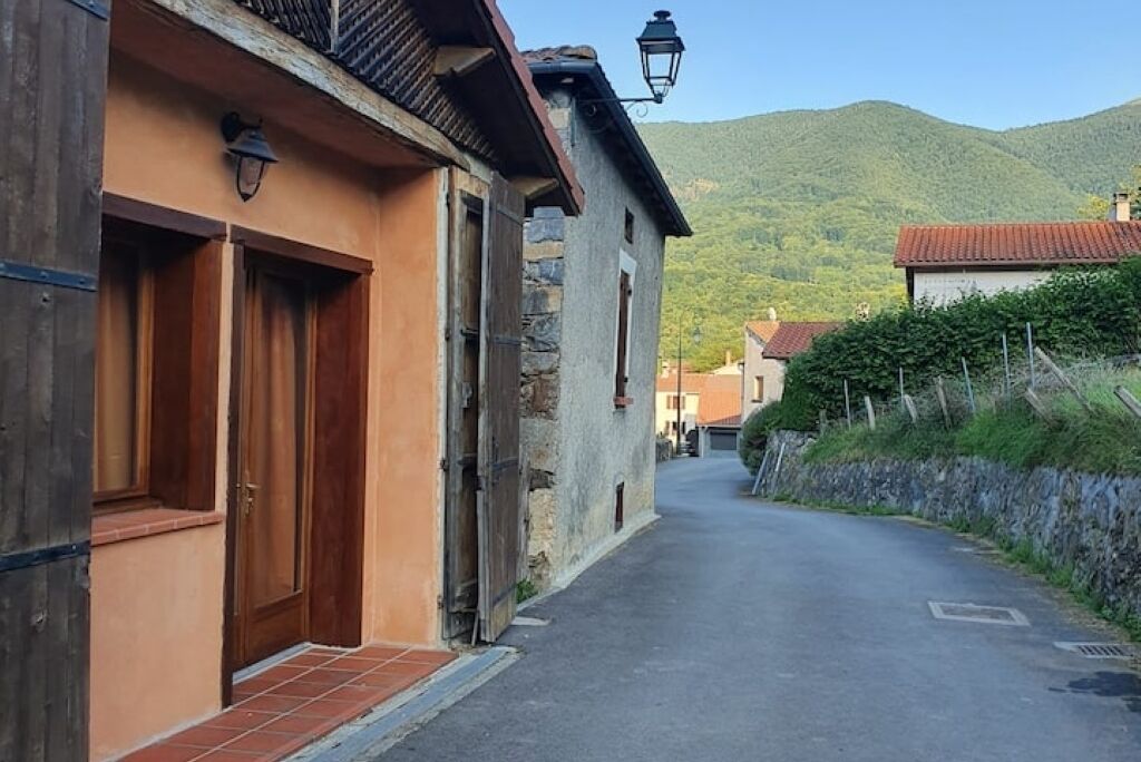 Una calle estrecha flanqueada por edificios, un muro de piedra a la derecha y una montaña al fondo.