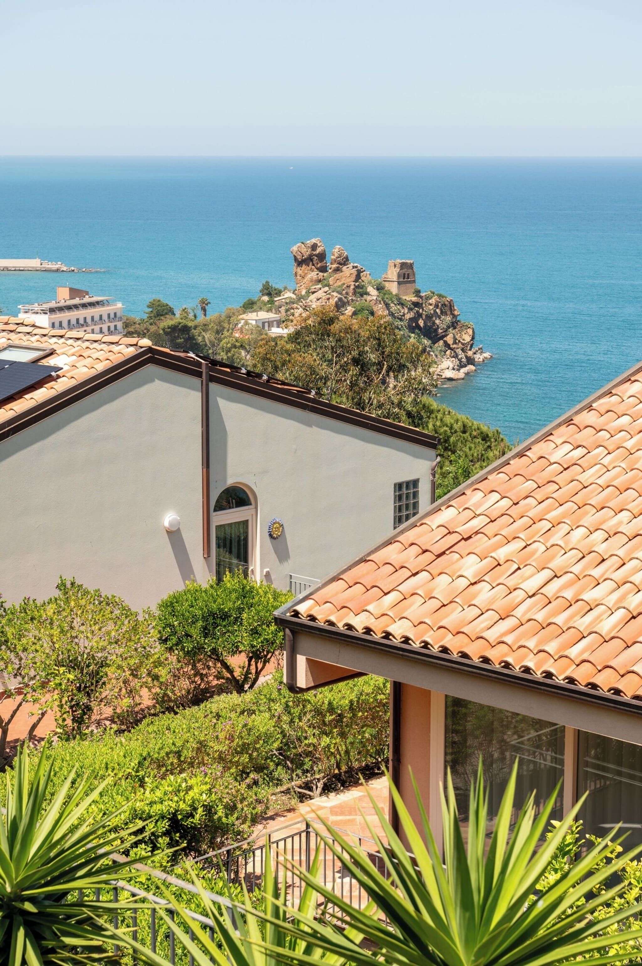 Un edificio costiero con tetto in tegole, vista sul mare e un promontorio roccioso sullo sfondo.