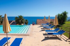 A poolside area with blue lounge chairs and umbrellas, a clear view of the ocean, and a tiled roof structure.