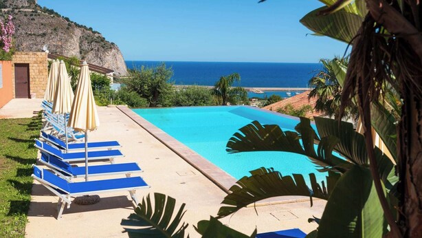 A pool area with blue lounge chairs and umbrellas, a clear view of the sea, and a mountainous backdrop.