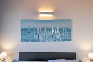 A bedroom with a bed, a wall-mounted light, and a painting of sailboats on the wall.