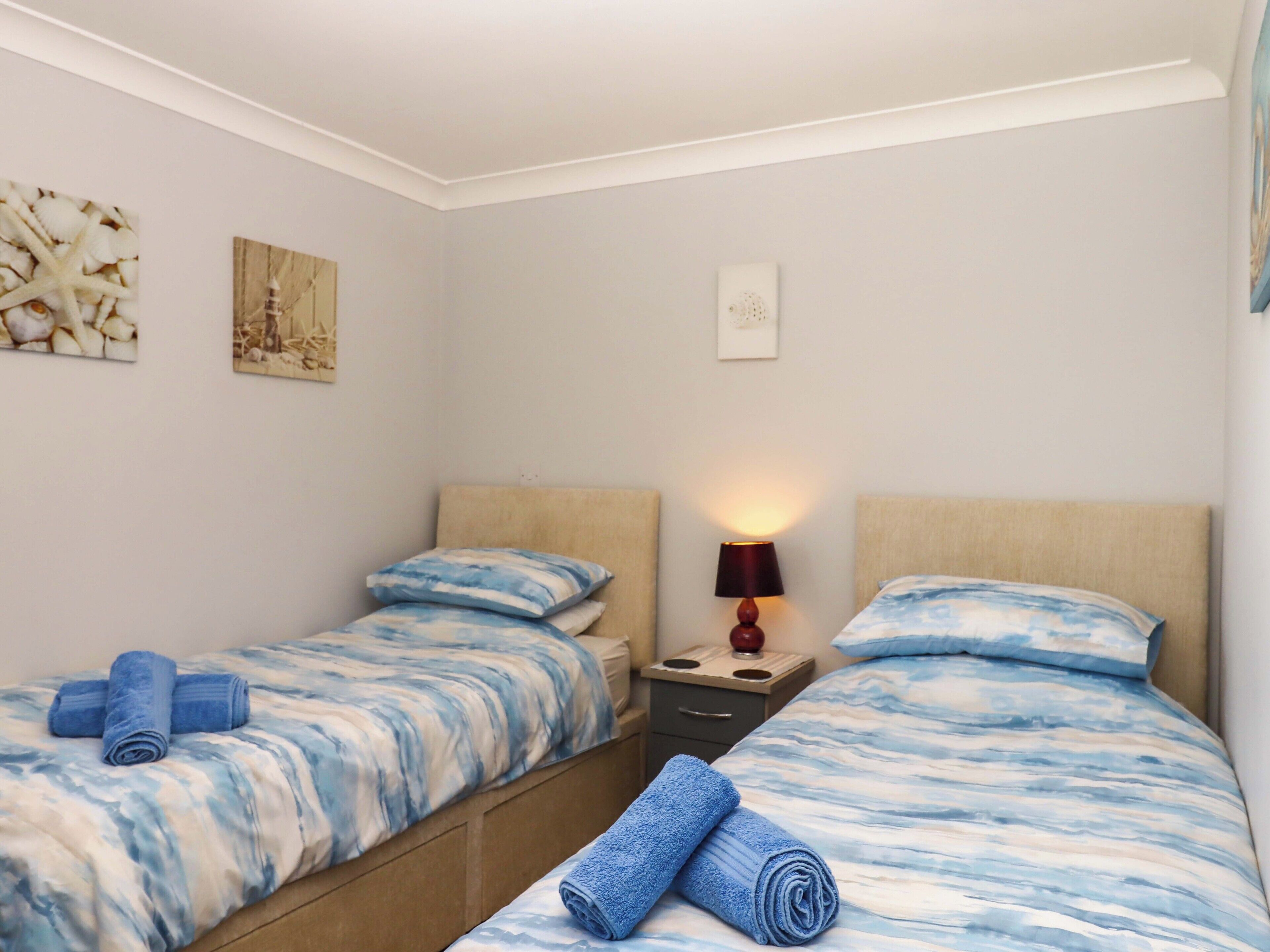 Habitación de hotel con dos camas individuales, cada una con colcha y almohadas de diseño azul y blanco. Hay una mesita de noche con lámpara entre las camas, y una pequeña obra de arte en la pared sobre la mesita.