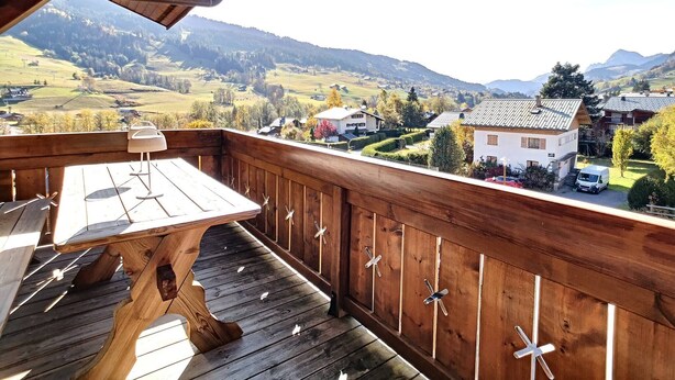 Eine Holzdar Terrasse mit Tisch und Stühlen, mit Blick auf ein malerisches Tal mit Häusern und Bergen.