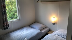 A small, single-room accommodation with two beds, a window with curtains, and a wall-mounted light fixture.