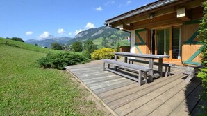 Eine Holzhütte mit einer Terrasse und einem Picknicktisch, umgeben von einer Wiese und Bergen im Hintergrund.