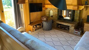 Ein gemütliches Blockhaushaus-Interieur mit Kamin aus Stein, einem an der Wand montierten Fernseher und einem großen Fenster mit Vorhängen.