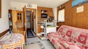 Un espace de vie compact comprenant un coin cuisine, un espace repas avec une table et des chaises, un canapé recouvert d’un tissu à motifs et une bibliothèque.