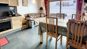 Un salon confortable avec un canapé, une table à manger et des chaises. Il y a une cuisine équipée d’une cuisinière et de placards. Une fenêtre offre une vue sur un paysage enneigé.