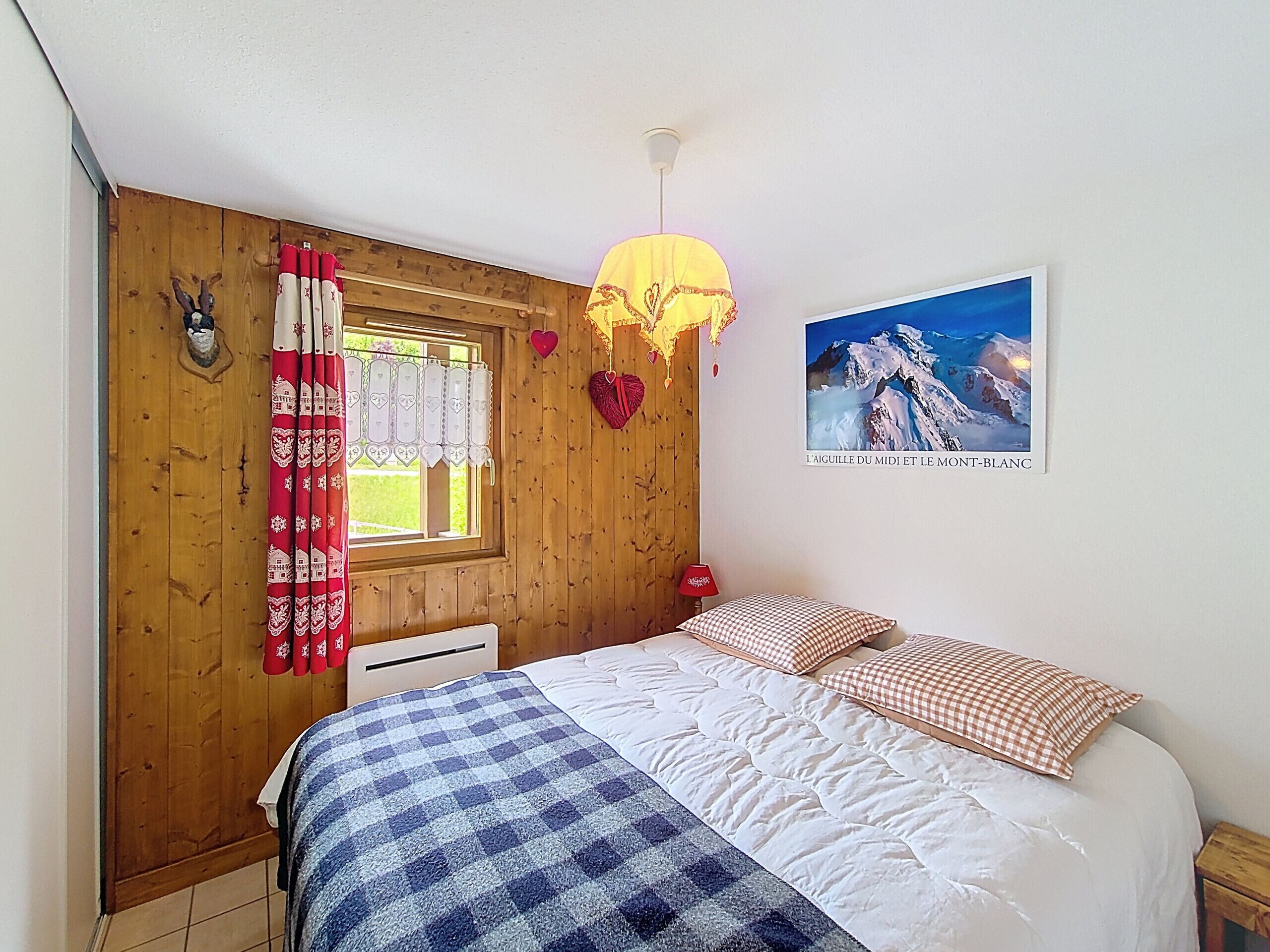 A bedroom with a wooden wall, a bed with checkered bedding, a window with curtains, a wall-mounted lamp, and a framed picture.