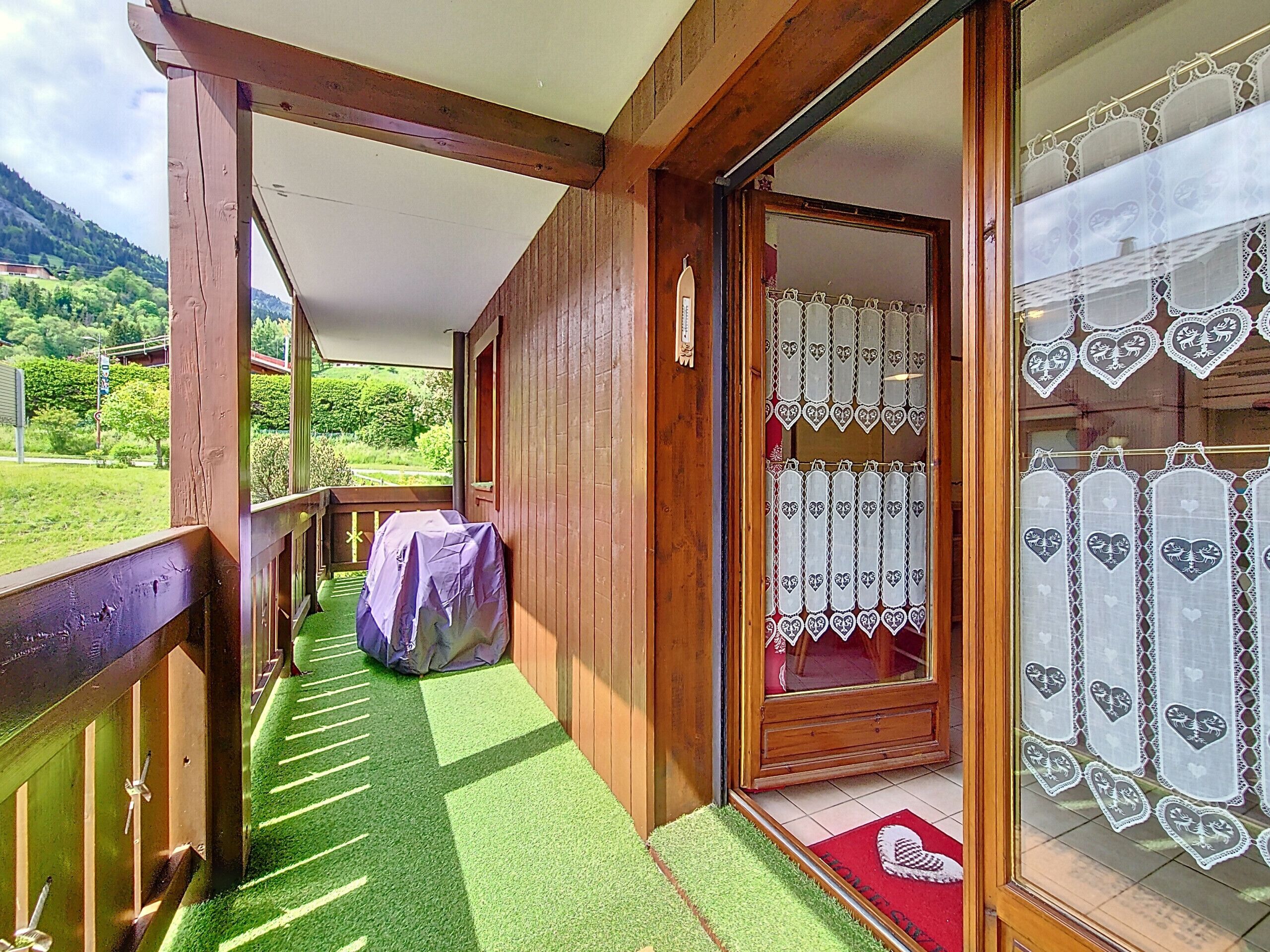 Uma varanda de madeira com vista para uma paisagem verde, uma área coberta com um tapete vermelho e uma porta com cortinas estampadas de corações.