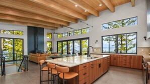 A modern kitchen with a large island, bar stools, and a view of the outdoors. - Cliff and Canopy - Lakefront Masterpiece w/Private Dock (Bonner County)