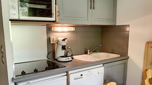 Een kleine keuken met een inbouwoven, een spoelbak en een compacte koelkast. Er is een houten tafel en een stoel.
