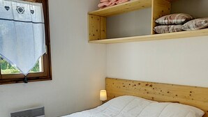 Een slaapkamer met een houten hoofdbord, een wit sprei, een bedlampje, een raam met gordijn en een plank met opgevouwen kleding.