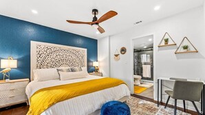 A modern bedroom with a large bed, a ceiling fan, a desk, and a bathroom visible through a glass door. - The Copper Cottage: An Eclectic Oasis with Hot Tub (Berkley)