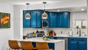 A modern kitchen with blue cabinets, a central island, and a dining area with brown chairs. - The Copper Cottage: An Eclectic Oasis with Hot Tub (Berkley)