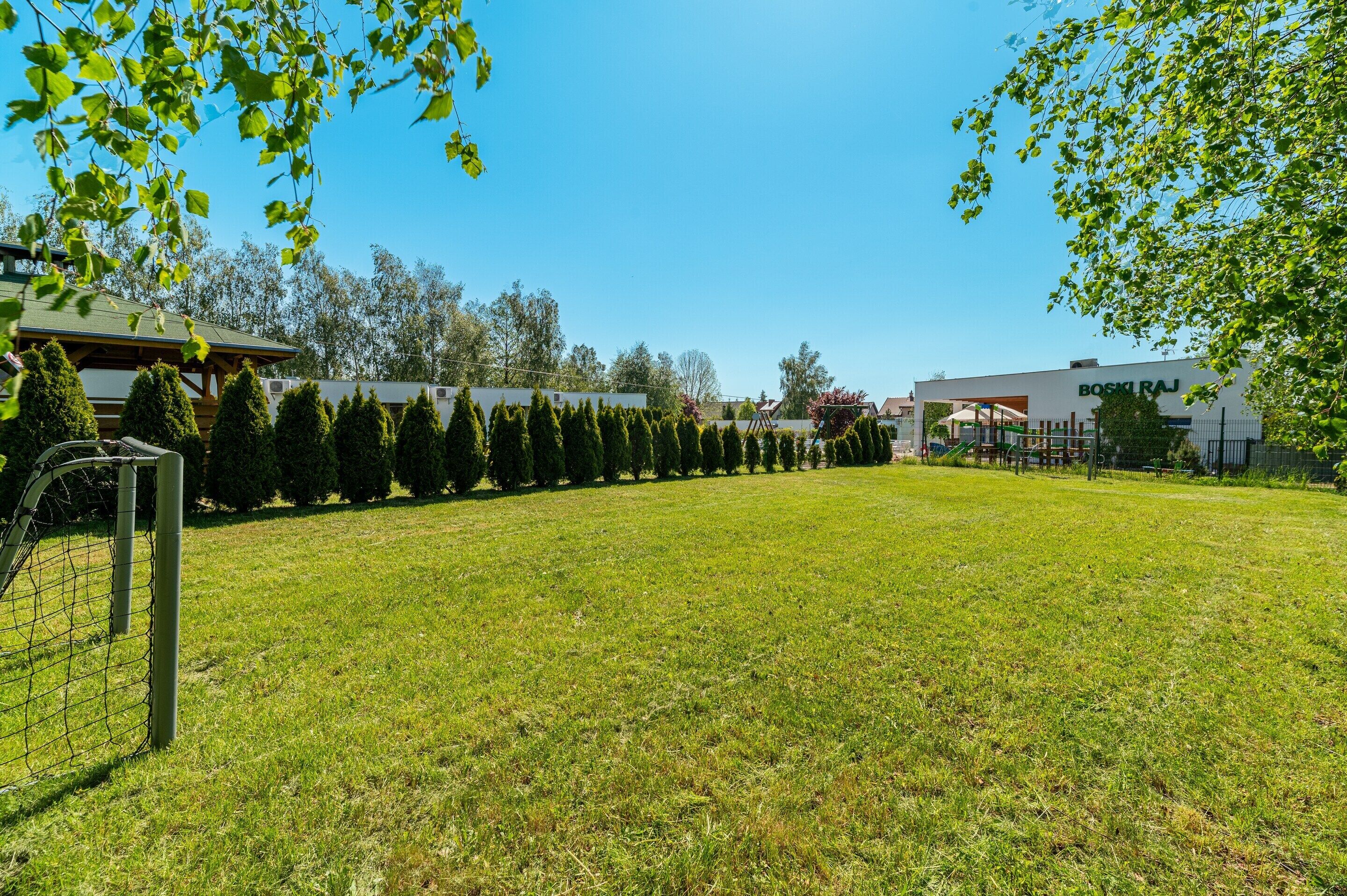 A grassy area with a fenced soccer goal, a row of neatly trimmed trees, and buildings in the background.