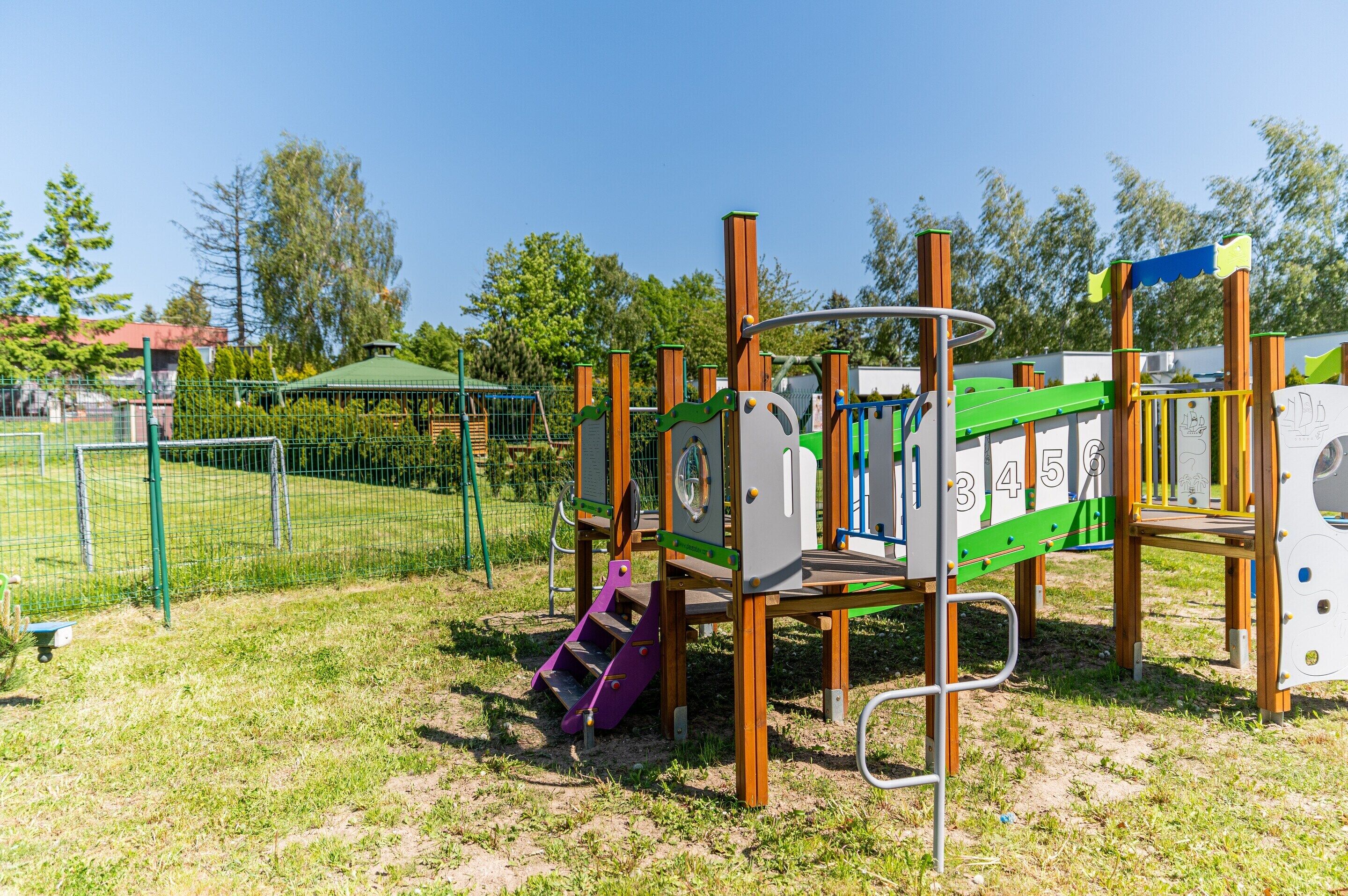 A playground with various play structures, including a slide and climbing elements, surrounded by a fenced area with grass and trees.