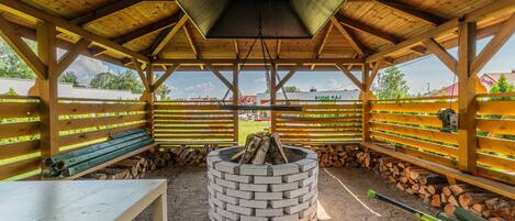 A covered outdoor area with a central fire pit, benches, and stacked firewood.